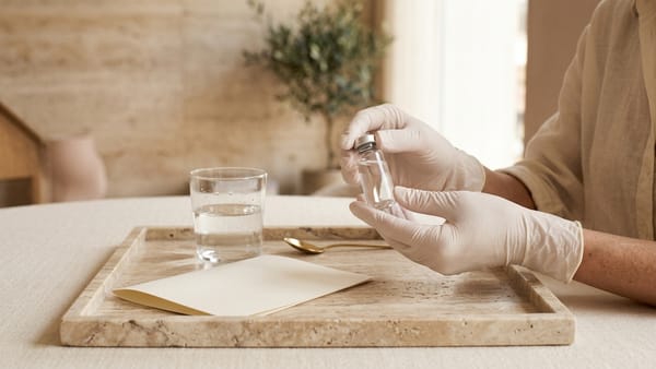 Mãos com luvas brancas a segurar um pequeno frasco de vidro sem rótulo sobre um tabuleiro de travertino, com um copo de água e uma folha em branco